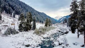 Scenic winter landscape in Pahalgam with snow-covered trees and mountains by a flowing river.